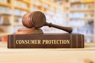 A wooden gavel rests on top of a thick book titled "CONSUMER PROTECTION," set against a blurred background of a library or bookshelf.