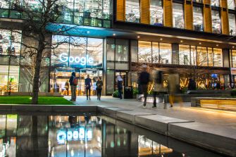 Exterior view of a Google office building with the Google logo and people walking outside near a reflecting pool.
