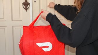 A DoorDash delivery person approaching a front door while carrying a red insulated delivery bag.