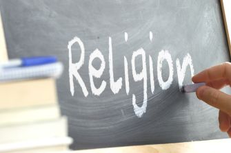 A hand writing "Religion" on a chalkboard, with books in the foreground.