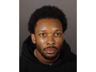 Mugshot of a man with short curly hair and a beard, wearing a black hoodie, looking upward against a gray backdrop.