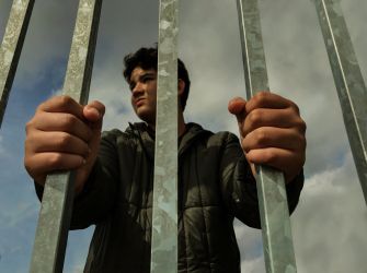 Person behind vertical metal bars, gripping the bars and looking outward.