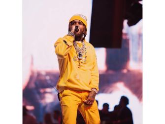 Male performer in a bright yellow outfit and beanie performing on stage with a microphone, blurred crowd in the background.