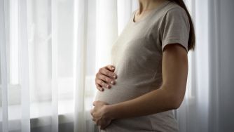 Side view of a pregnant woman in a gray shirt standing by a window with sheer white curtains, cradling her baby bump.