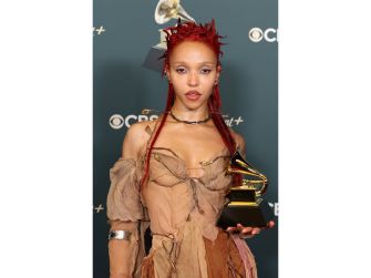 Woman with bright red, spiky hair wearing a distressed beige outfit, holding a trophy at an awards show with a logo-filled backdrop in the background.