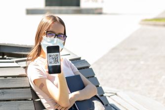 A young woman wearing a face mask holds a smartphone displaying a QR code while seated outdoors.