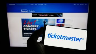 A person holding a smartphone displaying the Ticketmaster logo in front of a computer screen showcasing concert ticket promotions.