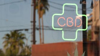 Neon sign displaying "CBD" in a green cross against a blurred outdoor background with palm trees.