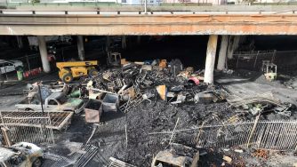 A burnt landscape under the freeway, showing damaged vehicles and debris from a pallet-fueled fire.
