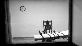 An execution chamber with a gurney and restraints, viewed through a window. A clock is visible on the wall.