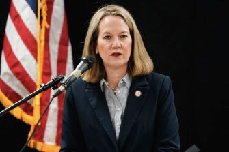 Blonde woman in a dark blazer speaking at a microphone, with an American flag in the background.