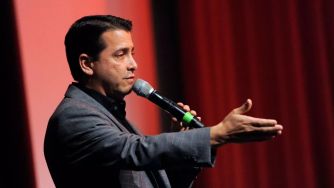 A man speaking into a microphone with his hand raised, set against a backdrop of red curtains.