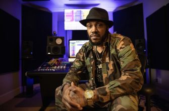 Man wearing camouflage clothing and a black hat seated in a recording studio with a mixing console, speakers, and a computer monitor in the background.