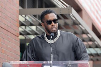 Sean Combs speaking at a public event, wearing sunglasses and jewelry, with a microphone in front of him.