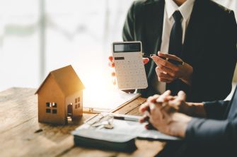 A person in a suit holding a calculator next to a small house model on a desk, with another person's hands visible in the foreground, indicating a discussion about home appraisal.