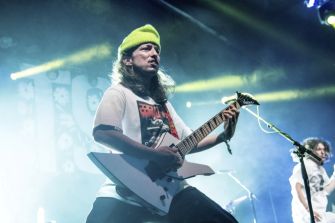 Guitarist wearing a yellow beanie playing a white flying V electric guitar on stage during a live concert, with another musician visible in the background.