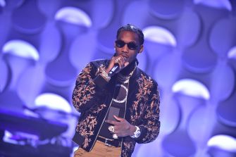 Offset performing on stage in a black-and-gold embroidered jacket and sunglasses, holding a microphone.