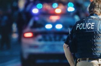 Police officer standing near a police car with flashing lights in a public setting.