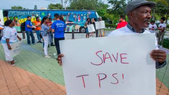 A man holds a sign reading "SAVE TPS!" at a rally advocating for the continuation of Temporary Protected Status for Haitian immigrants, while other supporters gather in the background.