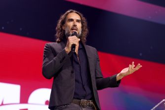 A man with long, wavy hair and a beard stands on a stage, speaking into a handheld microphone against a red and blue backdrop.