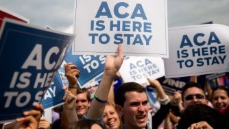 A diverse group of people holding signs reading "ACA IS HERE TO STAY" in support of the Affordable Care Act.