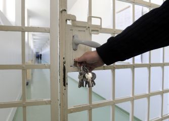 A hand holding keys prepares to unlock a prison gate, leading to a hallway.