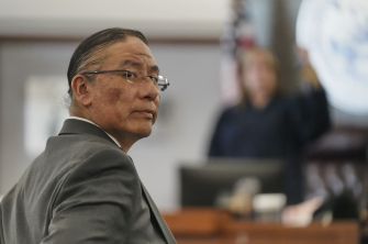 Side profile of a man in a suit wearing glasses in a courtroom, with blurred figures in the background.