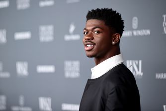 Close-up of a young man with short curly hair and a diamond earring, wearing a black and white suit, posing in front of a gray event backdrop.