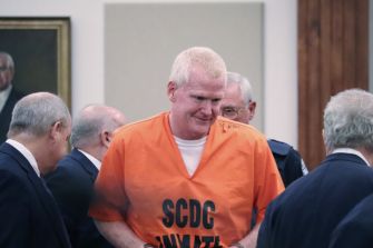 An inmate in an orange SCDC jumpsuit is escorted by officials in suits down a hallway.