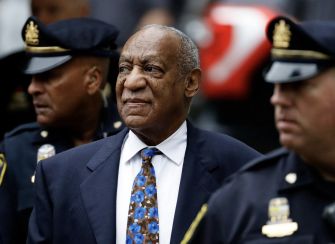 Older man in a dark suit and blue floral tie in a courtroom setting, with police officers in uniform nearby.