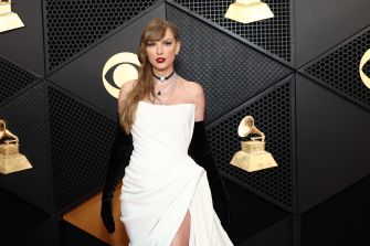 Celebrity on a red carpet wearing a white one-shoulder gown and black gloves, posing in front of a backdrop with gold gramophone logos.