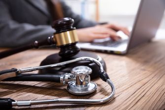 A gavel and stethoscope on a table, with a person using a laptop in the background, symbolizing medical malpractice law and legal processes.
