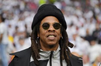 A man with long hair and sunglasses smiles while wearing a black hat and jacket, surrounded by an audience in the background.