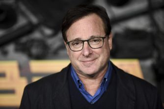 A smiling man wearing glasses and a dark jacket, standing in front of a colorful background.