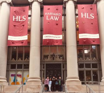Group of people on the steps of Harvard Law School, with red banners reading HLS and Harvard Law Orientation hanging between columns.