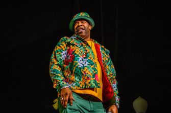 Male rapper on stage wearing a bright, multicolored patterned jacket and matching hat with jewelry against a dark background.