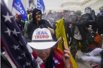 Protest outside the Capitol with American flags and police in riot gear; a person wearing a Team Trump hat stands in the foreground.
