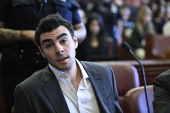 Young man in a suit sits in a courtroom during a federal hearing, with a microphone in front and blurred spectators in the background.
