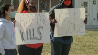 Students holding signs at a protest with messages about the dangers of hazing, including "HAZING KILLS" and "R.I.P. STONE."