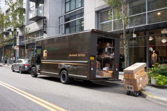 A brown UPS delivery truck with its rear doors open, showing boxes inside, parked on a city street; a dolly loaded with boxes sits on the curb beside a storefront.