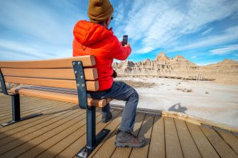 A person sitting on a bench in a national park, taking a photo with a smartphone against a scenic backdrop of rock formations.
