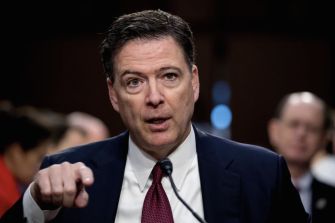 Close-up of James Comey in a dark suit and red tie, speaking into a microphone at a hearing.