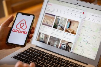 Close-up of a hand holding an iPhone with the Airbnb logo beside a laptop screen displaying the Airbnb website and a map.