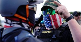 A member of a group wearing tactical gear and a patterned mask, engaged in a discussion with another individual, during a protest event.