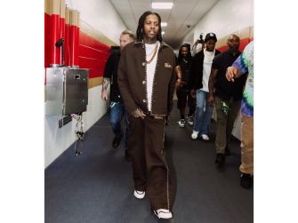 Man in a brown coordinated outfit walking down a hallway, flanked by an entourage and photographers in the background.