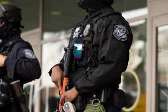 Two masked federal officers in black tactical gear with patches, standing outside a building.