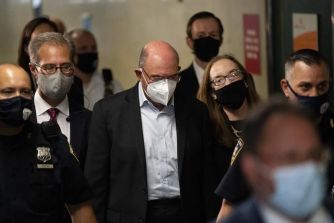 Group of formally dressed individuals wearing face masks walking through a hallway; a bald man in a dark suit at the center.