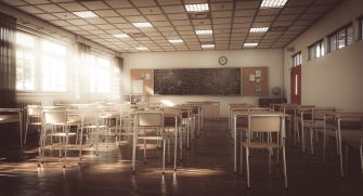 An empty classroom with wooden desks arranged in rows, sunlight streaming through the windows, and a chalkboard at the front.