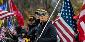 Rally scene with people holding American flags; a man wearing sunglasses and a cap stands in the foreground.