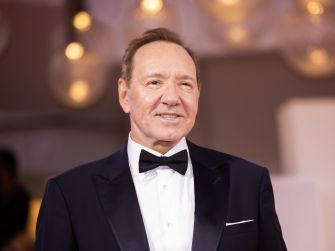 Close-up of a man in a black tuxedo and bow tie, smiling at a formal event with blurred warm lights in the background.